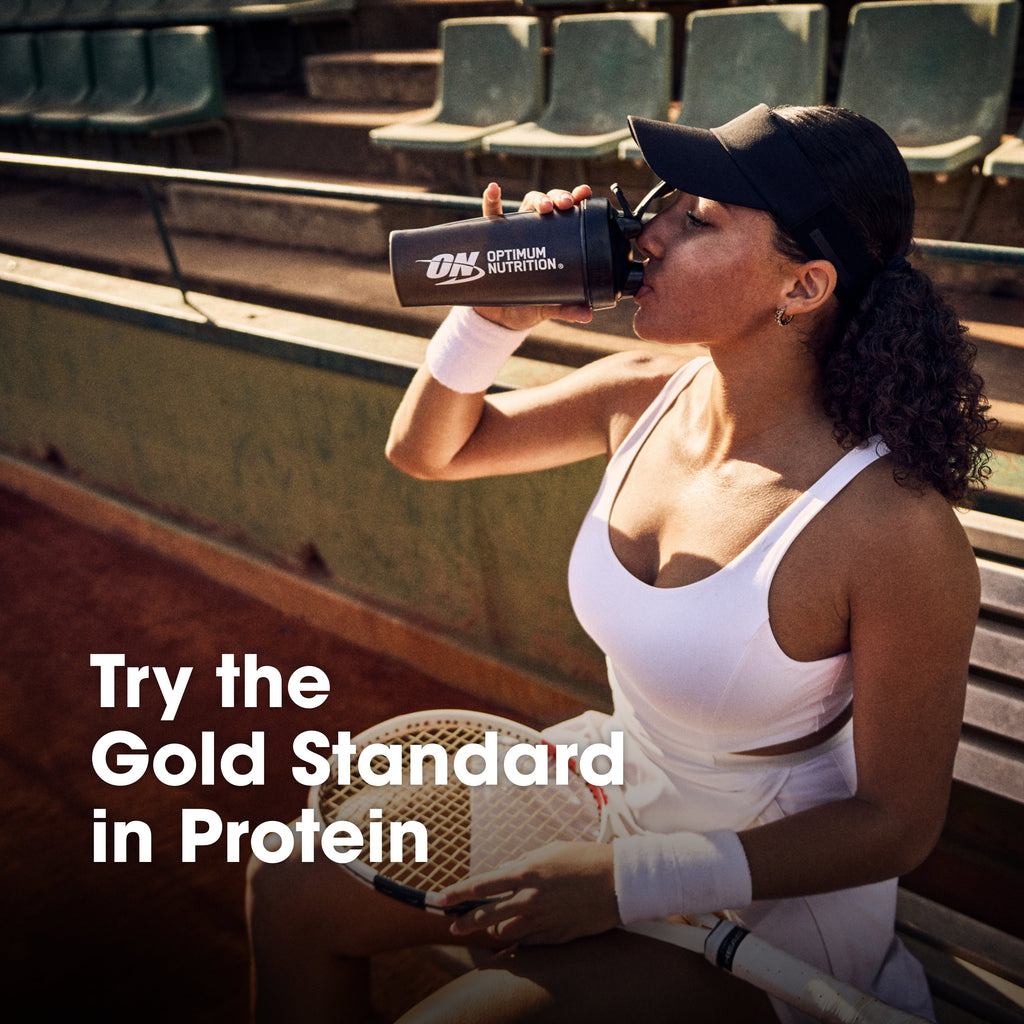 A woman in a white tennis outfit sits on stadium seating holding a protein shaker bottle with the Optimum Nutrition logo. Large text on the image reads 'Try the Gold Standard in Protein'.